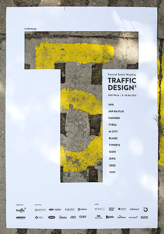 Plakat Traffic Design z wyciętą literą "T". Przez dziurę w plakacie widać różne tła: mur, graffitti, szklane szybki. 
